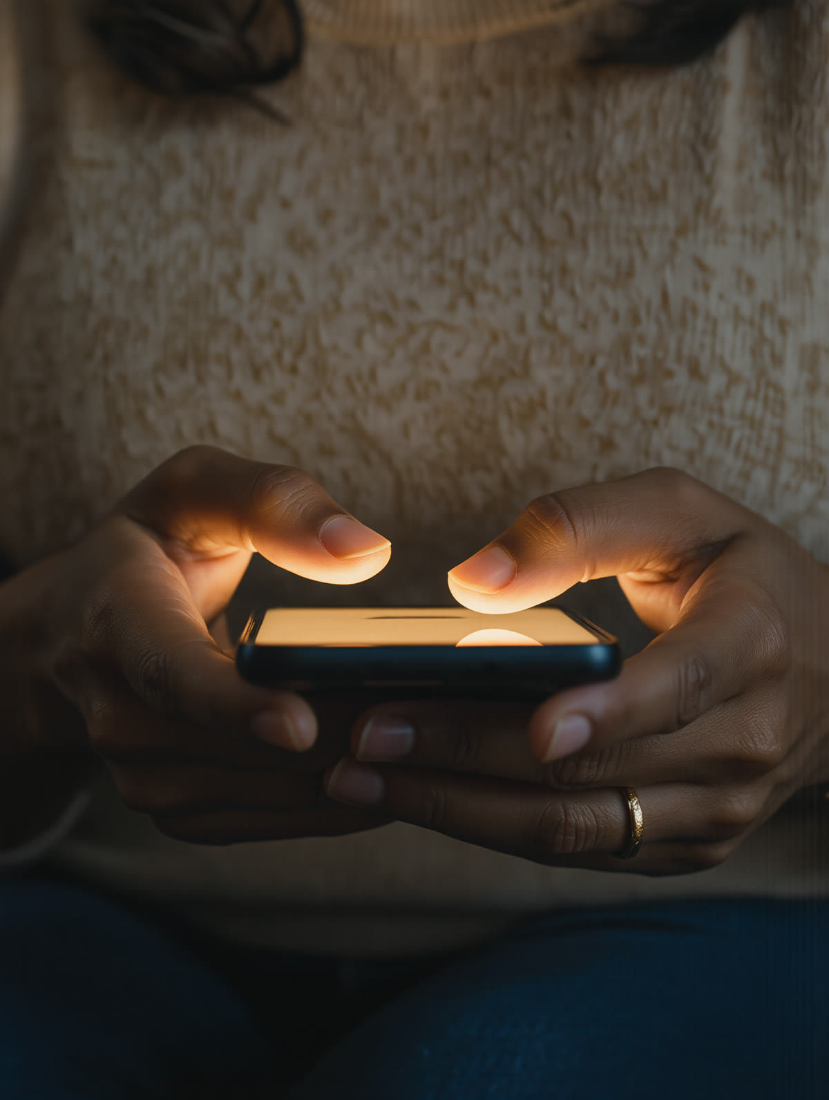 Hands hold a phone with a soft warm glow on its screen in a dim living room.